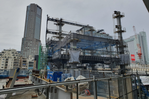 Großer Bauplatz in einer Stadt mit einem Metallzaun, Gebäuden, Pfählen, einer Plane, Fahrzeugen, Drähten und einem bewölkten Himmel, was den Bau eines neuen Kraftwerks anzeigt.