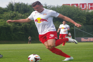 Ein professioneller Fußballspieler, der einen Ball auf einem grünen Feld kickt, mit "RB Leipzig" im Hintergrund unter einem klaren blauen Himmel.