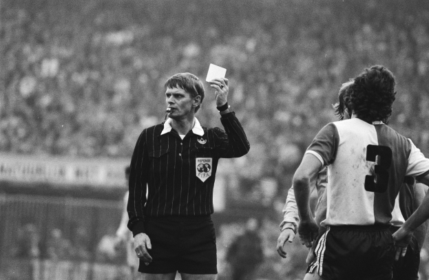 Schwarzes und weißes Bild eines Schiedsrichters mit einer roten Karte vor einer Menge von Menschen in Fußballuniformen, mit einem Zaun im Hintergrund.