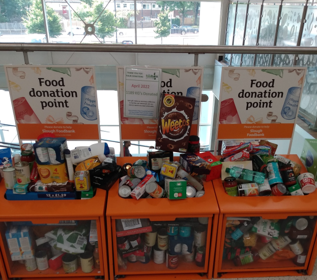 Orange Schränke mit Lebensmitteln wie Flaschen, Dosen und Kartons, neben Texttafeln, mit Fahrzeugen, Bäumen, Gebäuden und Himmel durch Glasfenster sichtbar.