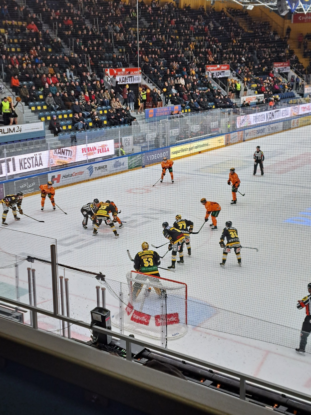 Gruppe von Menschen, die Hockey auf einem Eisstadion mit Netz und Umzäunung spielen, unter Stadionbeleuchtung, mit Zuschauern im Hintergrund und Schildern