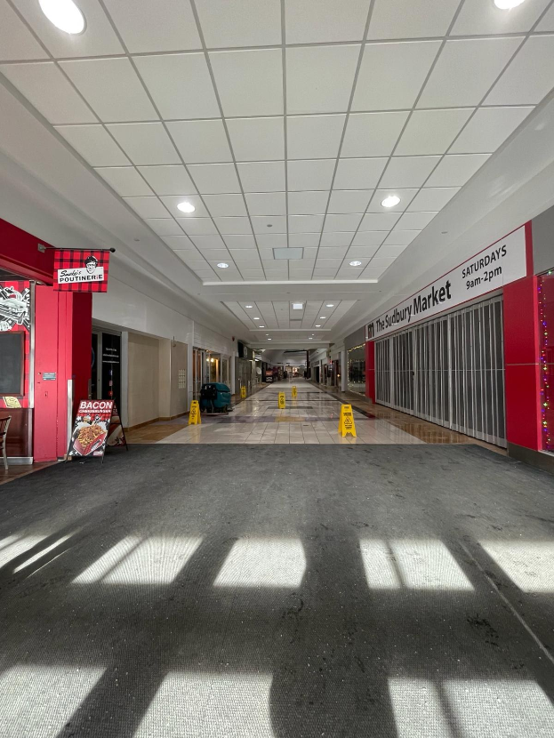 Das Innere eines Einkaufszentrums mit Deckenbeleuchtung, einem Stuhl und einer Tafel, auf der links 'Sydbury Market - Vorher und Nachher' steht, sowie verstreuten Gegenständen rechts und auf dem Boden.