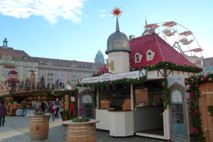 Ein geschäftiger Weihnachtsmarkt in Nürnberg, Deutschland mit Menschen um geschmückte Stände, festliche Lichter, Schmuck, Gebäude, ein Riesenrad und einen bewölkten Himmel, mit einer Tafel mit Text auf der rechten Seite.