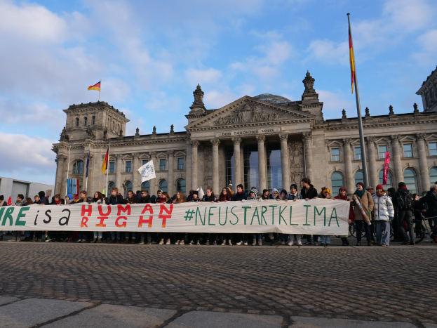 Eine Gruppe von Menschen hält ein Transparent mit der Aufschrift "Wir sind ein Menschenrecht" vor dem Reichstaggebäude in Berlin, Deutschland, mit seinen architektonischen Details und umgeben von Fahnen unter einem bewölkten Himmel.