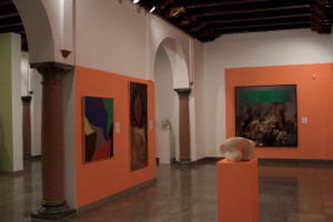 Ein Ausstellungsraum voller verschiedener Kunstwerke, darunter Gemälde, Skulpturen und eine Statue, mit Deckenleuchten und Säulen, die die Ausstellung 'Emanationen: Die Kunst der kameralosen Fotografie' in der Govett-Brewster Art Gallery zeigen.