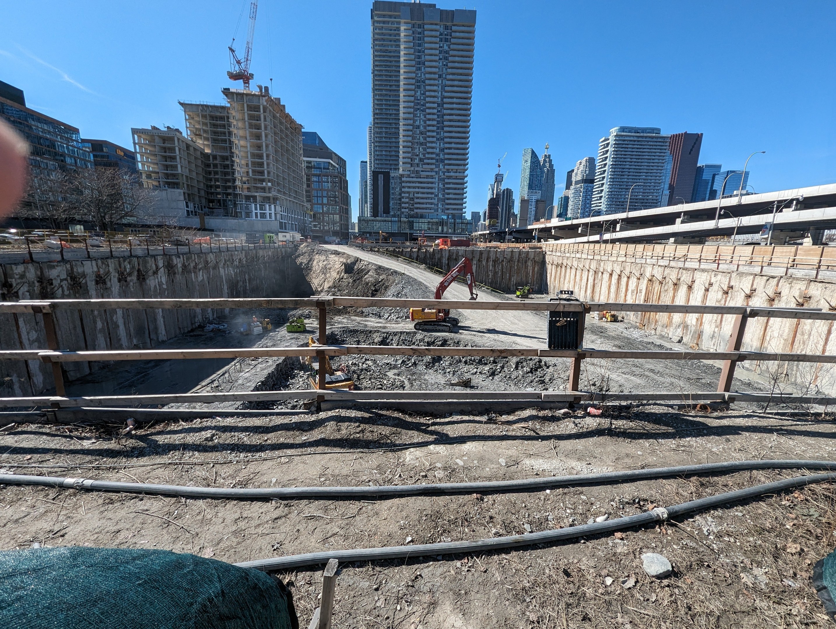 Eine Baustelle für einen neuen Toronto-Wasserfront-Park mit einem großen Haufen Erde, umgeben von Gebäuden, Bäumen, Pfählen, Lampen, Geländern, Fahrzeugen und anderen Gegenständen, mit einem sichtbaren Himmel im Hintergrund und einer Hand eines Menschen auf der linken Seite.