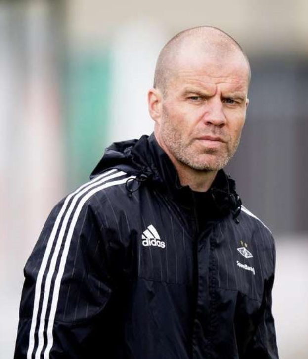 Baldiger Trainer in einem blauen Jackett mit weißen Streifen und weißem Kragen, der auf einem Fußballfeld steht und nachdenklich die Hände gefaltet hat.