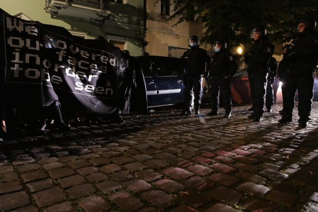 Eine Gruppe von Polizisten in Helmen steht auf einer Kopfsteinpflasterstraße bei Nacht, mit einem Banner haltend, mit Fahrzeugen, Gebäuden, Bäumen und Beleuchtung im Hintergrund.