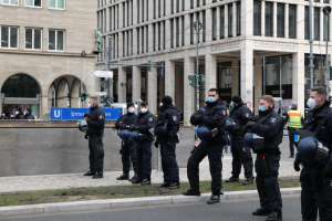 Eine Gruppe von Polizisten in schwarzen Uniformen und Masken steht vor einem Gebäude mit Glasfenstern und Säulen in Berlin, Deutschland, einige halten Helme, mit Laternen, Verkehrszeichen, Tafeln mit Text, einer Statue, anderen Menschen und einer grasbewachsenen Fläche im Hintergrund.