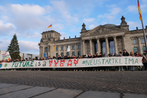 Eine Gruppe von Menschen hält ein Transparent mit der Aufschrift 'Zukunft ist ein Menschenrecht' vor dem Reichstag in Berlin, umgeben von Bäumen, Fahnenmasten und einem bewölkten Himmel.