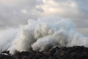 Eine große Welle schlägt gegen die Felsen am Strand, mit Wasser, das gegen den Strand schwappt und einer bewölkten Kulisse.