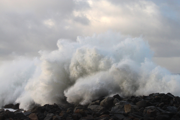 Eine große Welle schlägt gegen die Felsen am Strand, mit Wasser, das gegen den Strand schwappt und einer bewölkten Kulisse.