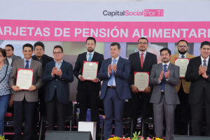 Gruppe von Menschen auf einer Bühne, die klatschen und Zertifikate halten, mit Stühlen in einem Halbkreis angeordnet, einem Banner mit der Aufschrift "Capital Social for Ti" im Hintergrund und Blumensträußen und Lautsprechern unten.