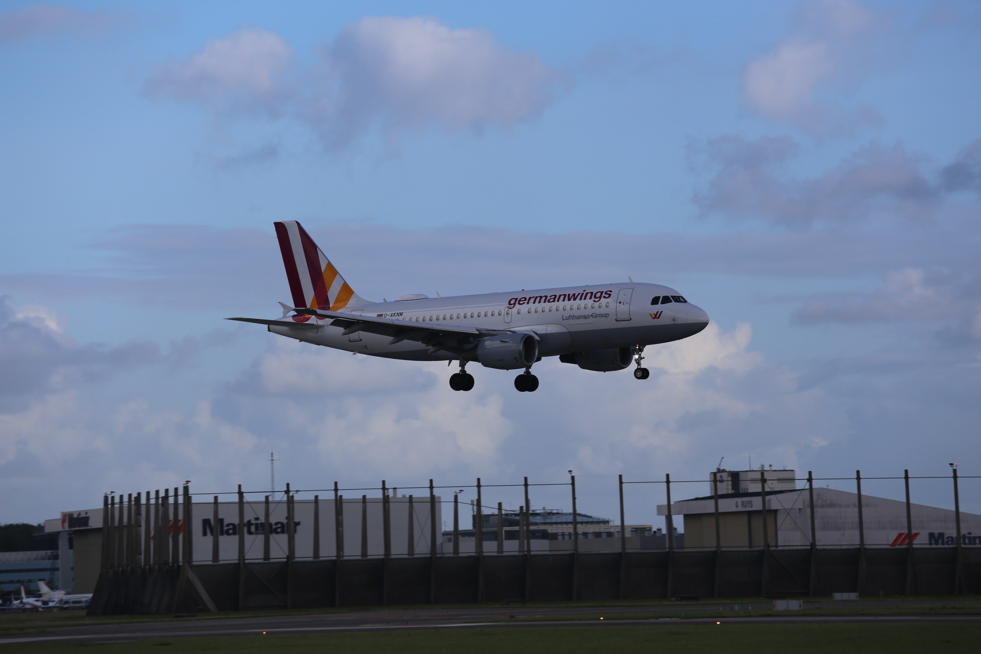 Germanwings Airbus A320-200 beim Start vom Frankfurt Airport mit Gras darunter, einem Zaun im Vordergrund, Gebäuden und Bäumen im Hintergrund und einer bewölkten Himmel