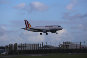 Germanwings Airbus A320-200 beim Start vom Frankfurt Airport mit Gras darunter, einem Zaun im Vordergrund, Gebäuden und Bäumen im Hintergrund und einer bewölkten Himmel