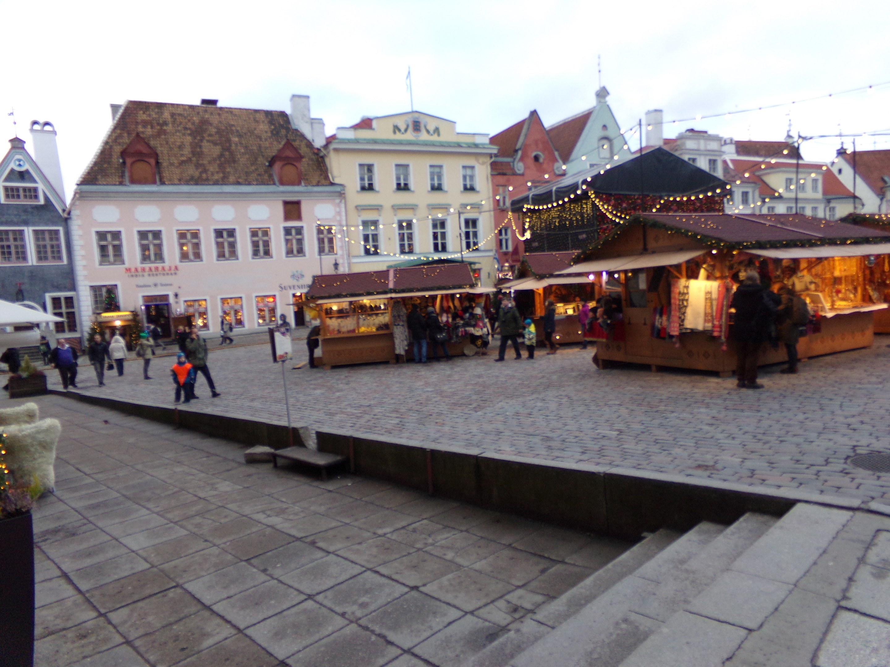 Ein geschäftiger Weihnachtsmarkt in Tallinn, Estland, mit Menschen um geschmückte Stände, festlicher Beleuchtung und Topfpflanzen, vor Wolkenhimmel und Gebäuden mit beleuchteten Fenstern.