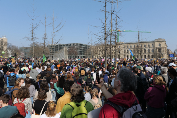 Eine große Menschenmenge mit Schildern und Taschen steht vor einem Gebäude mit Fenstern, Bäumen und einem klaren Himmel, was auf eine Klimaprotest demonstration hinweist.