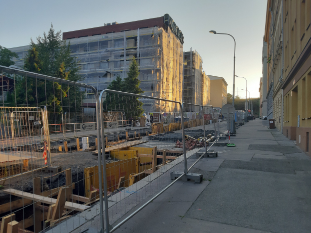 Stadtstraße mit einer Baustelle in der Mitte, umgeben von Gebäuden, Straßenlaternen, Straßenlaternen, Fahrzeugen, Absperrpollern, Bäumen und einem klaren blauen Himmel.