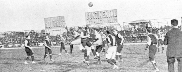 Ein Schwarz-Weiß-Bild einer Gruppe von Männern, die auf einem Feld Fußball spielen, mit einem Ball in der Luft und einer Zuschauermenge im Hintergrund, mit Tafeln, die wahrscheinlich textbezogen zur Partie sind.