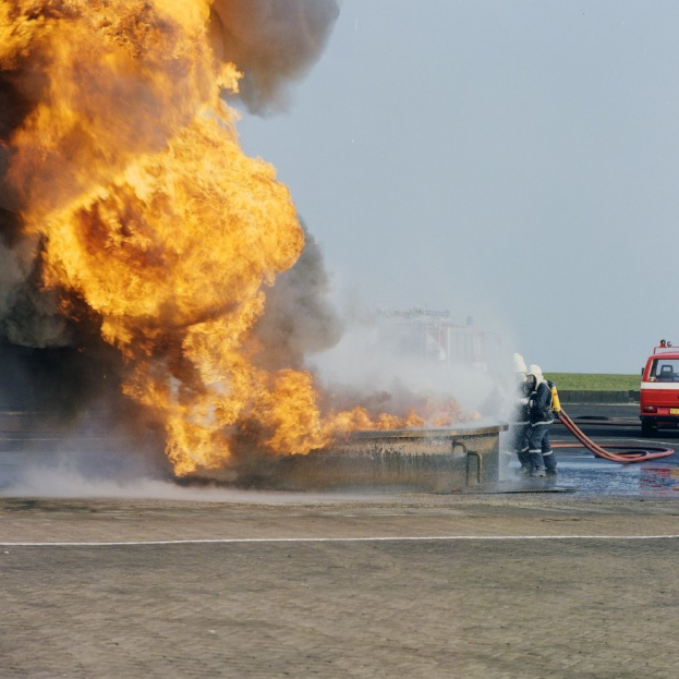 Ein Feuerwehrauto steht in Flammen an der Seite einer Straße, mit zwei Personen in Helmen und Schlöschen in der Nähe, einem Fahrzeug und dem Himmel im Hintergrund.