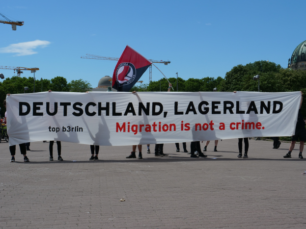 Eine Gruppe von Menschen hält ein Transparent mit der Aufschrift "Deutschland, Lagerland Migration ist kein Verbrechen" und eine Flagge und steht auf dem Boden mit Straßenlaternen, Bäumen, Kränen, einem Gebäude und einem bewölkten Himmel im Hintergrund.