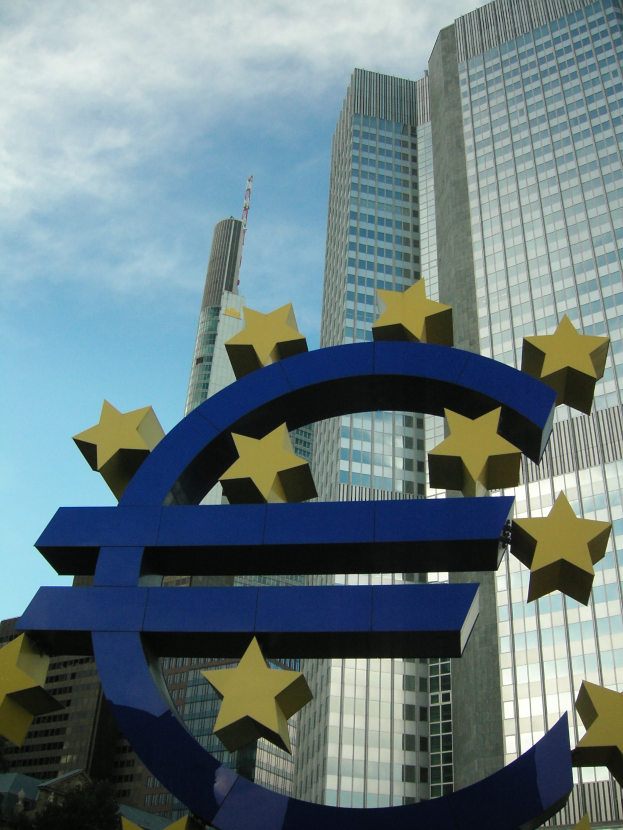 Das Euro-Zeichen steht prominent vor dem Hauptquartier der Europäischen Zentralbank, einem modernen Glasgebäude, das von Bäumen umgeben ist und unter einem bewölkten Himmel liegt.