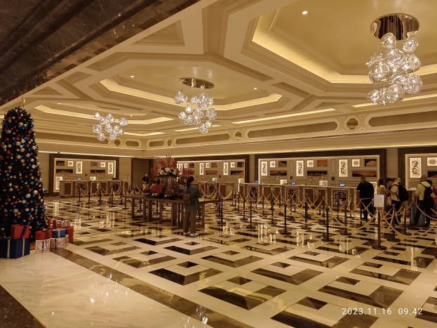 Eine gro\sse Lobby mit einem zentralen Weihnachtsbaum, umgeben von Geschenkboxen und festlicher Dekoration, mit Menschen, Absperrgittern, Tischen mit Blumenbouquets und gerahmten Fotos an den Wänden, unter einer Decke mit Lichtern und Kristalll"ustern.