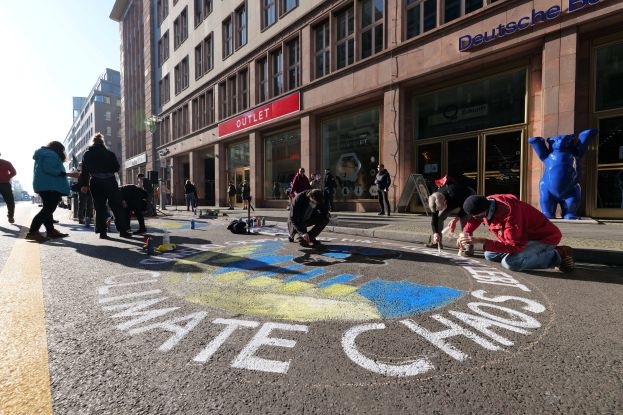 Menschen sitzen vor einem Gebäude mit Fenstern und Namensschildern auf dem Boden, umgeben von Flaschen und anderen Gegenständen, während sie an einer Klimawandel-Demonstration in Berlin teilnehmen, die von Bäumen und einem klaren blauen Himmel umgeben ist.