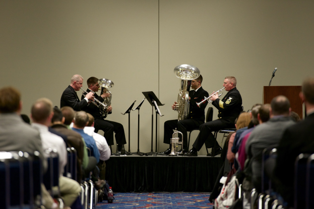 Eine Gruppe von Männern, die auf einer Bühne Brass-Instrumente spielen, mit Notenständern, einem Podium mit Mikrofon und einem Publikum, das auf Stühlen sitzt.