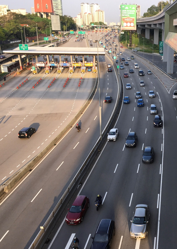 Eine vielbefahrene Autobahn mit zahlreichen Autos und Motorrädern, Verkehrsleitkegeln, Hinweistafeln, Laternenmasten, einer Brücke, Bäumen, Gebäuden und einem klaren blauen Himmel.