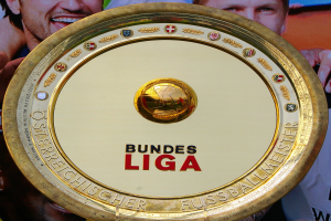 Ein Mann hält einen goldenen Schild mit "Bundesliga" darauf, mit einem Banner im Hintergrund mit drei Personen.