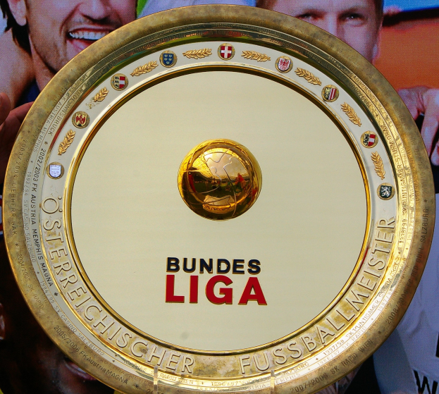 Ein Mann hält einen goldenen Schild mit "Bundesliga" darauf, mit einem Banner im Hintergrund mit drei Personen.