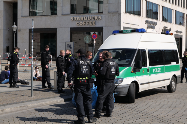Eine Gruppe von Polizisten steht vor einem Starbucks-Café, rechts davon parkt ein Van und links stehen und sitzen ein paar Menschen; im Hintergrund sind ein Gebäude, ein Schild, ein Laternenmast und ein Zaun zu sehen.