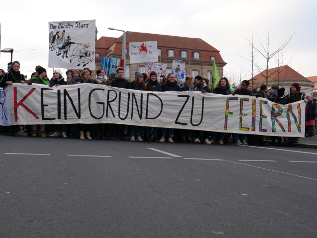Gruppe von Menschen mit einem Transparent "Kein Grund zu Feiern" bei einer Demonstration gegen Sparmaßnahmen der deutschen Regierung, mit Straßeninfrastruktur und Gebäuden im Hintergrund bei klarem Himmel.