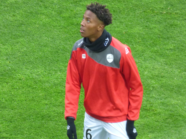 Ein junger Mann in einer roten Jacke und weißen Shorts steht auf einem Fußballfeld und trägt Baseballhandschuhe, während er entschlossen nach rechts schaut.