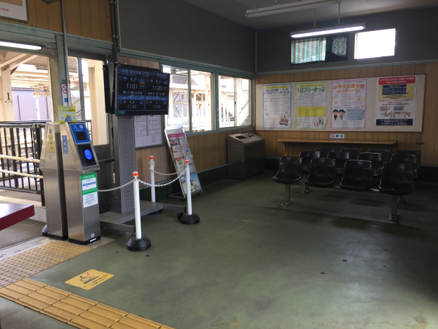Ein belebter Bahnhofseingang mit Sitzgelegenheiten, einem Service-Schalter, Informationsanzeigen, Plakaten, Deckenleuchten, Geländern und Ticketautomaten.