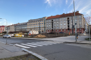 Baustelle für ein neues Wohngebäude vor dem Hintergrund einer städtischen Straße mit parkenden Autos, Laternen, Bäumen, Gebäuden und einem bewölkten Himmel.