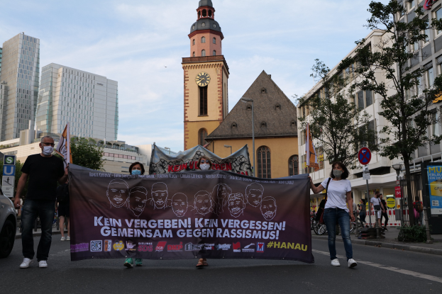 Eine Gruppe von Menschen mit Masken, die eine Straße entlanggehen und ein Transparent halten, mit einem geparkten Auto auf der linken Seite, Gebäuden, Bäumen, Schildern, Polen, einem Uhrenturm und einem klaren blauen Himmel im Hintergrund.