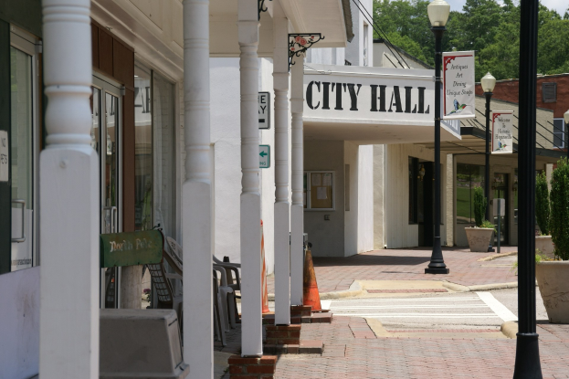 Ein Rathaus umgeben von Gebäuden, Straßenmasten, Straßenlaternen, Namenschildern, Kraftfahrzeugen, Absperrgittern, Stühlen, Topfpflanzen, Bäumen und einem bewölkten Himmel.