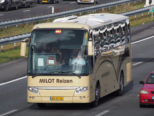 Ein Minibus und ein rotes Auto fahren auf einer Autobahn mit Leitplanken und Gras an den Seiten und Texttafeln im Hintergrund.