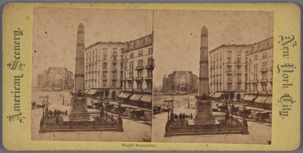 Altes Schwarz-Weiß-Foto einer Stadtstraße mit einem zentralen Denkmal, flankiert von Gebäuden, Straßenmöbeln, parkenden Autos und Fußgängern auf dem Gehweg unter einem sichtbaren Himmel.