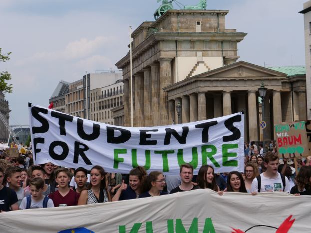 Schüler marschieren in Berlin mit einem leuchtend bunten "Students for Future"-Schild vor einem Hintergrund aus Gebäuden, Bäumen und Himmel.