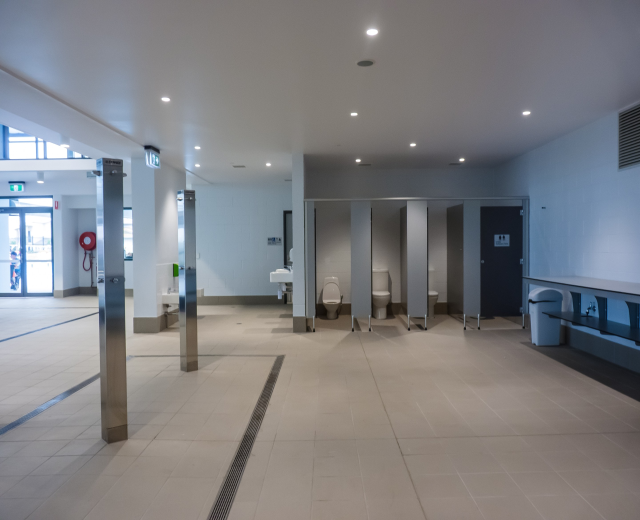 Große Öffentliche Toilette mit mehreren Toiletten, tragenden Säulen, einem Mülleimer, einer Tür, einem Schild, einem angebrachten Feuerlöscher und Deckenlampen auf dem Dach.